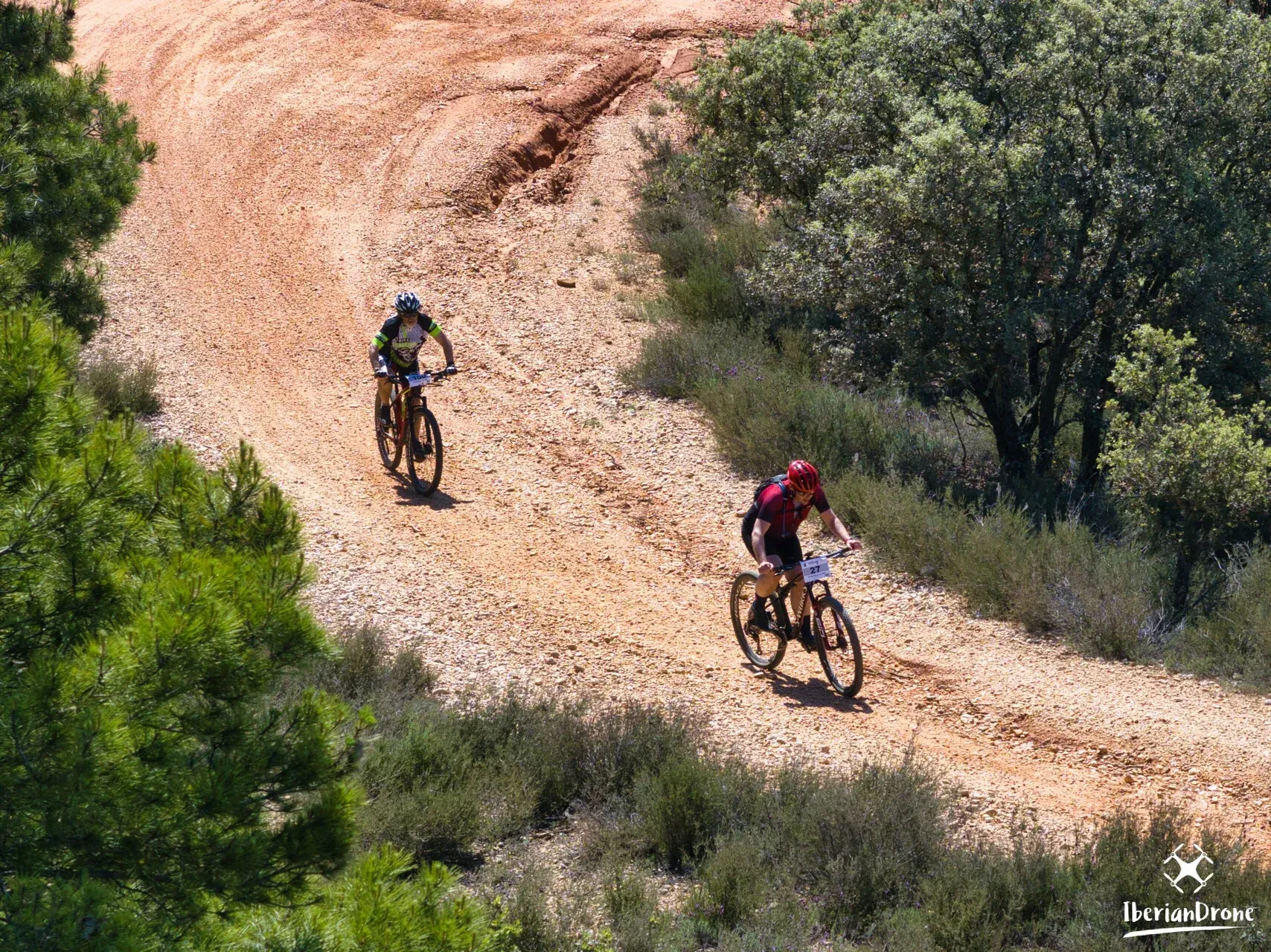 Dos participantes trazan una curva de tierra roja entre pinos y monte bajo.